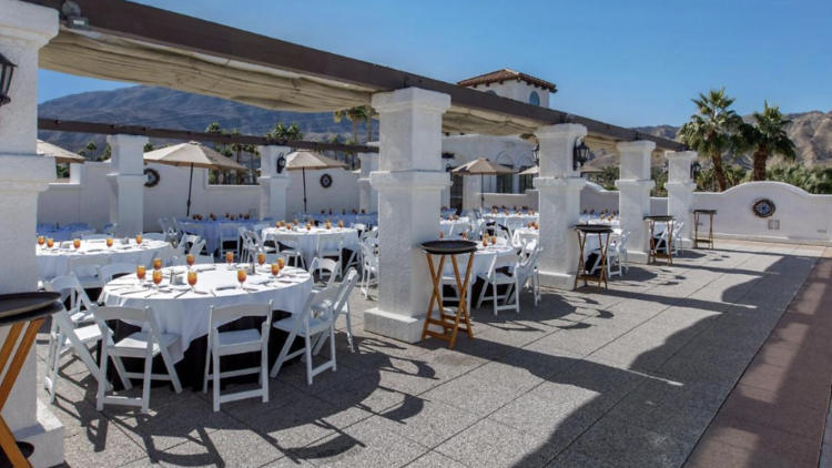 Sunny terrace with a row of dining tables filled with wine glasses.