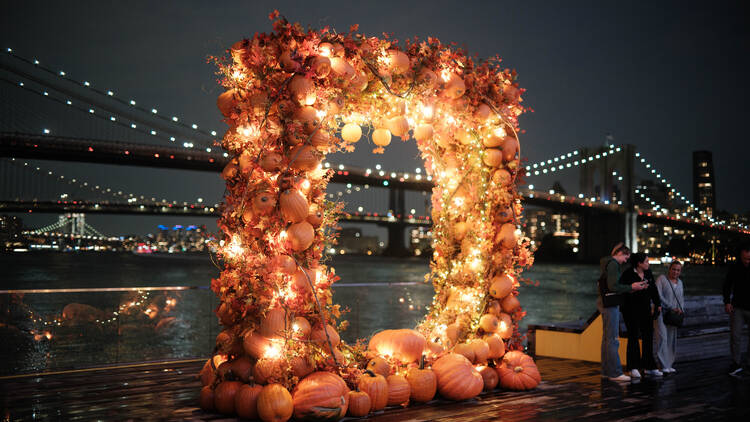 A pumpkin arch with lights and the Brooklyn Bridge in the background.