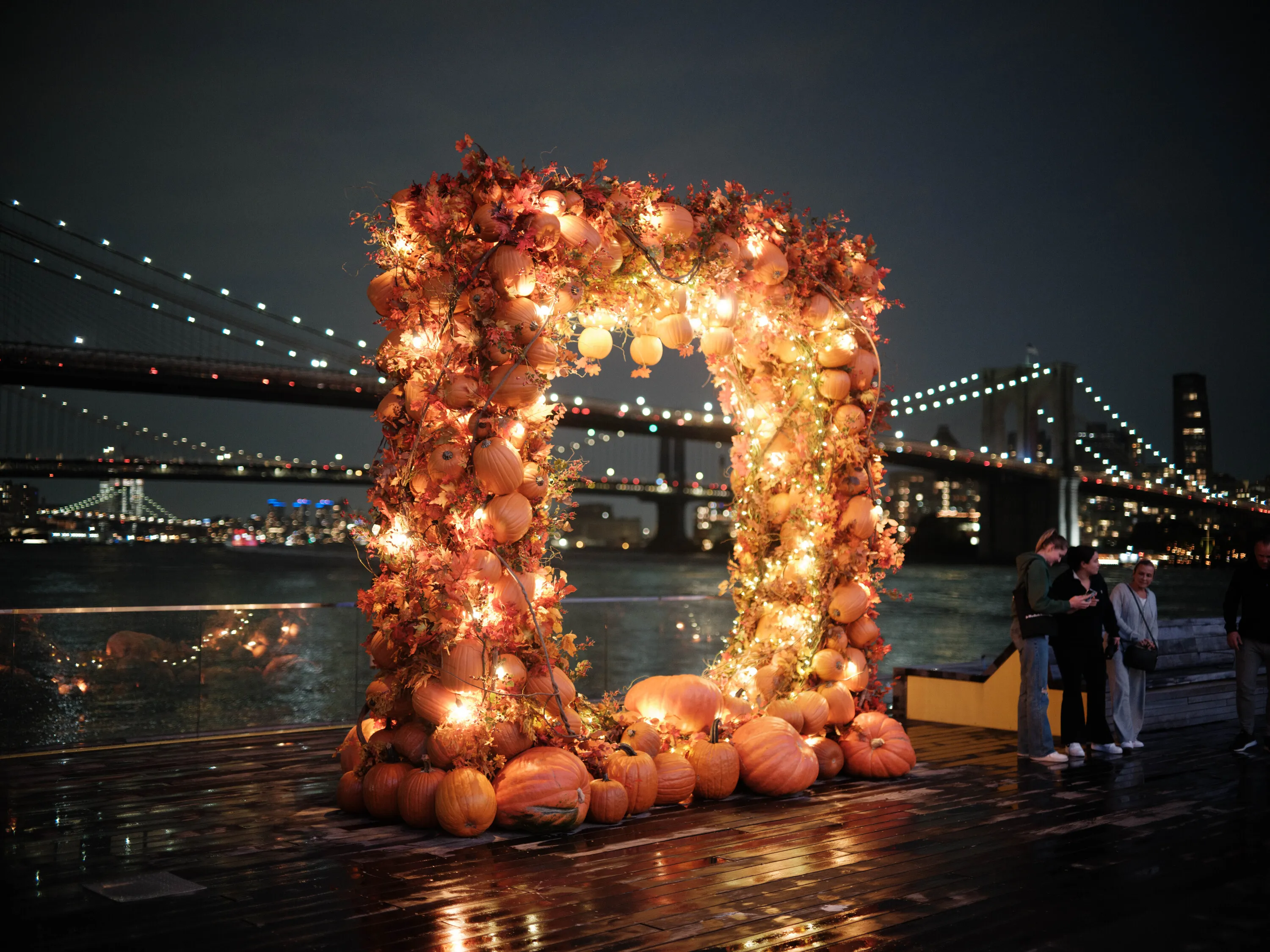 A pumpkin arch with lights and the Brooklyn Bridge in the background.