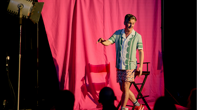 Ash Flanders on stage in front of a pink fabric backdrop wearing a colourful top and shorts