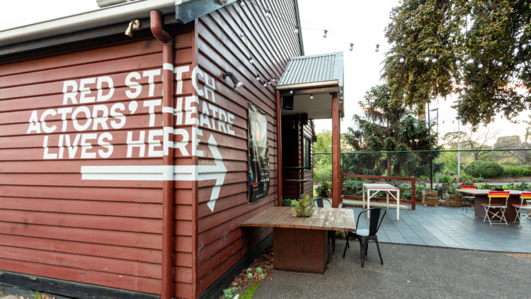 A side view of the dark red weatherboard building that houses Red Stitch and a small courtyard