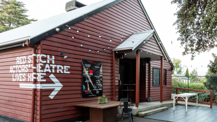 A side view of the dark red weatherboard building that houses Red Stitch and a small courtyard