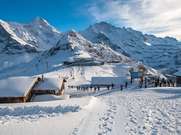 Lots of people with snowboards and skis on the mountain top getting ready for snow fun. Sun shining/sunlight on the snow covered wooden cottages, in Engelberg, Mount Titlis, Switzerland.