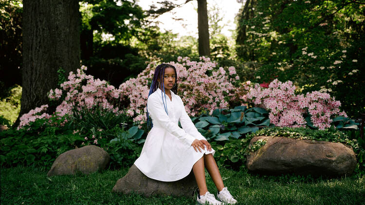 A portrait of a young Black girl in a white dress and sneakers with flowers and trees behind her.