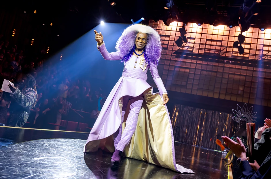 A man in a purple costume and wig walks on stage.