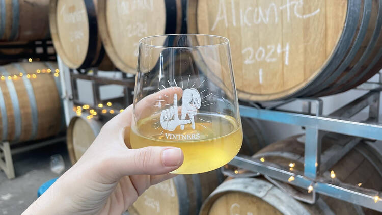 A glass of wine held up in front of barrels of Los Angeles wine.