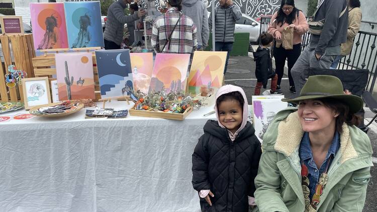 A woman and child pose in front of a table of artwork at Handmade Market Collective.