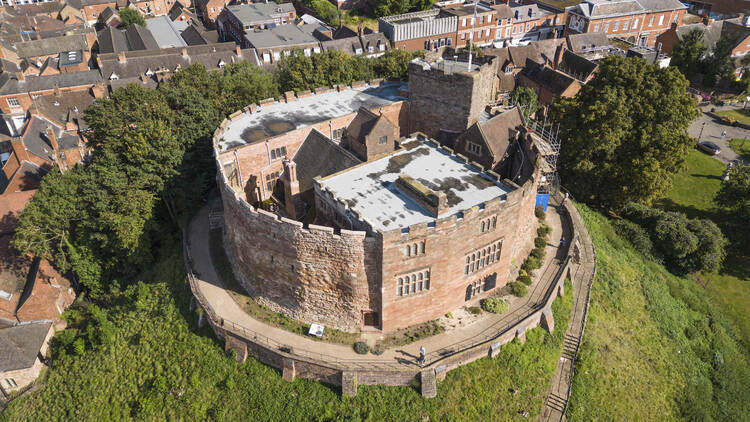 A photograph of Tamworth Castle, showing an old stone structure on a green hill with scaffolding.