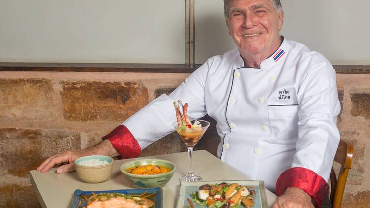 Chef David Zisman veste uniforme e senta à mesa com vários pratos tailandeses.