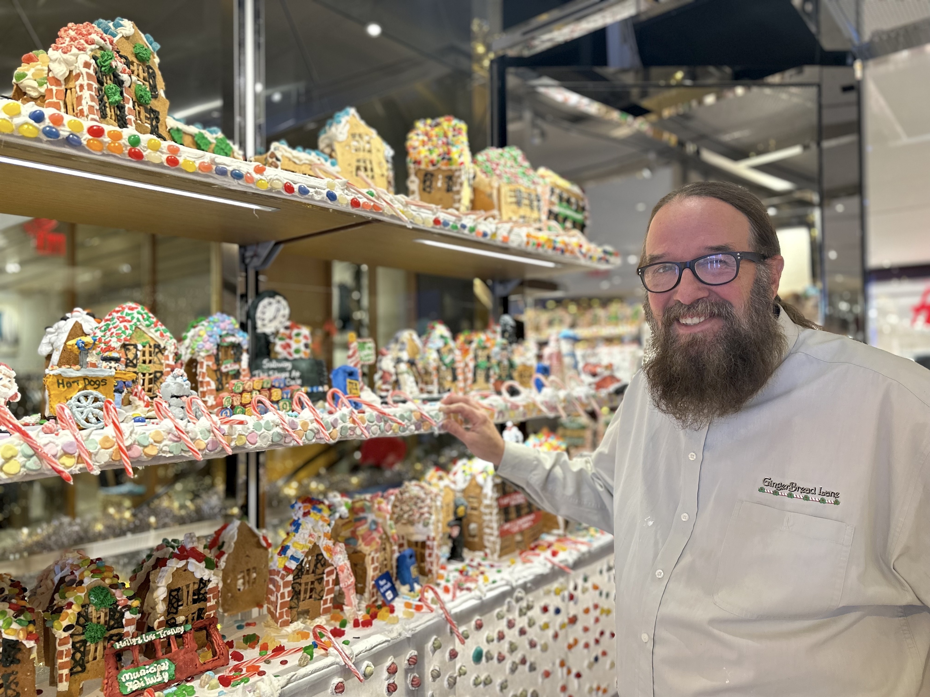 A man in a gray collared shirt stands next to a gingerbread village.