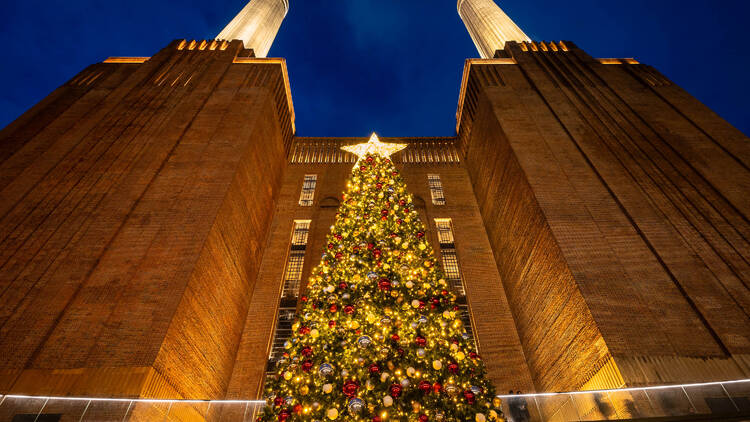 Battersea Power Station's two chimneys viewed from below during darkness, with a 40ft Christmas tree decorated with warm yellow lights and red baubles in between them