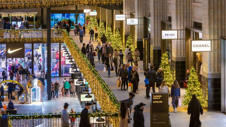 Shoppers walking past individual fir trees decorated with gold lights lining the walkways of Battersea Power Station's turbine hall