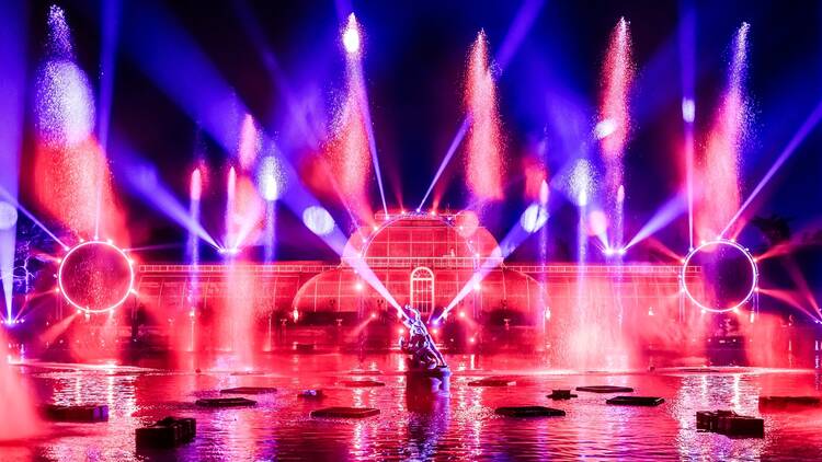 The Palm House at Kew Gardens lit up with red and blue lasers, viewed from across a lake, with a statue of Hercules wrestling a snake in the centre