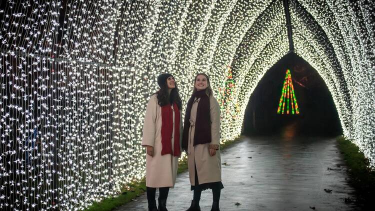 Two women dressed in warm coats, hats and scarves stand in the centre of a tunnel constructed of white fairy lights