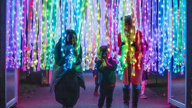 A family of three pose beneath a mass of colourful ribbons decorated with fairy lights