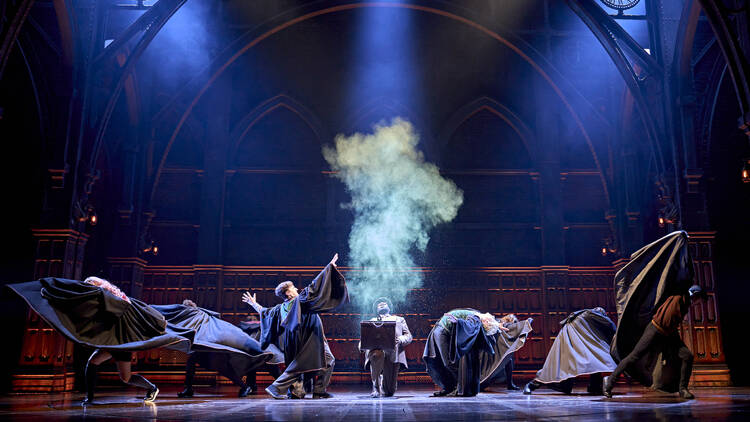 A production shot of Harry Potter: The Cursed Child, with a spell being cast by a boy with wand raised, and green smoke rising from a briefcase held by a kneeling man