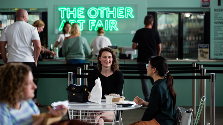 People sit eating food in front of a neon sign reading the other art fair