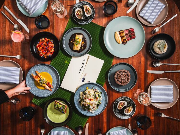 A spread of dishes, including oysters, steak and dips, on a dark wood table with peoples' hands outstretched to dine 