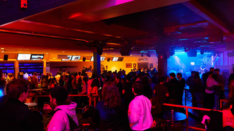 A crowd dancing illuminated by purple and blue light, with a DJ booth in the right corner