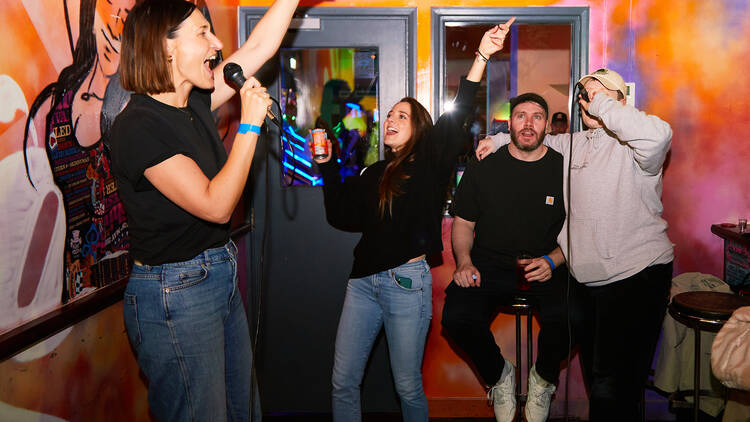 Four people standing in a graffiti-decorated karaoke booth, two singing into microphones