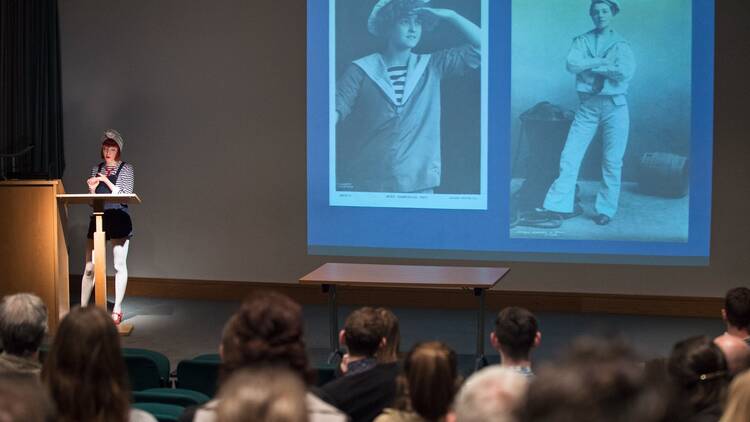 A woman dressed as a sailor stands at a lectern in front of an audience, with two black and white photographs of sailors projected onto the wall behind her