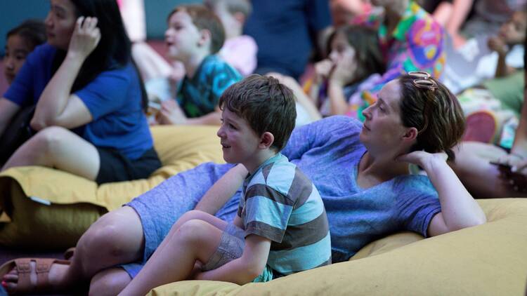 An adult and a child sitting on a beanbag watching a performance. 