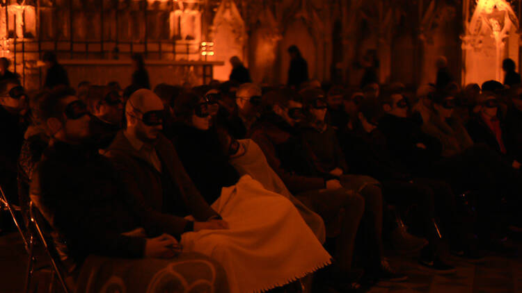 An audience sitting on chairs, all wearing blindfolds, in a dimly lit room