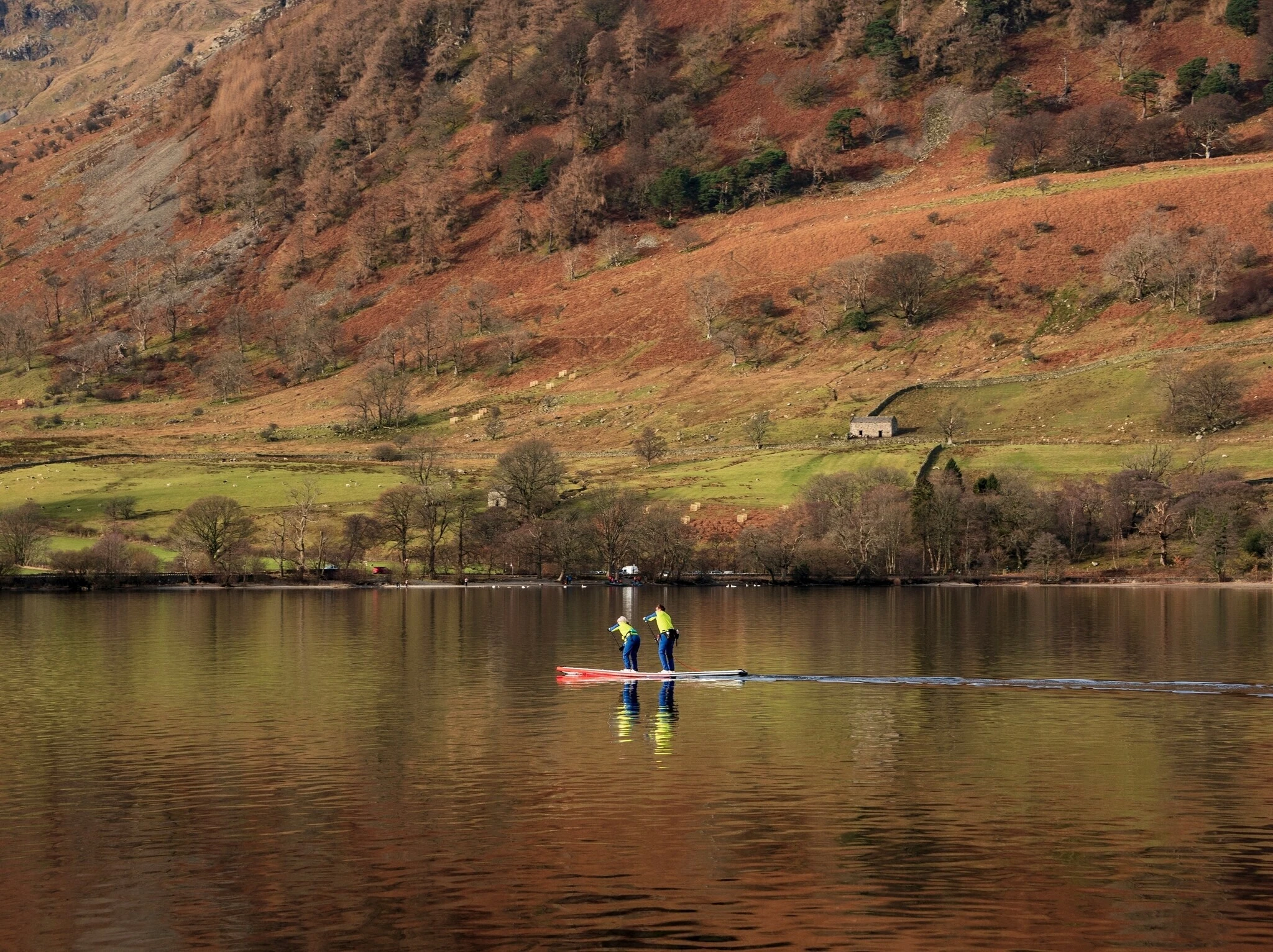 Beautiful Winter landscape views of mountain ranges around Ullswater in Lake District viewed from boat on lake with unidentified people paddle boarding on lake