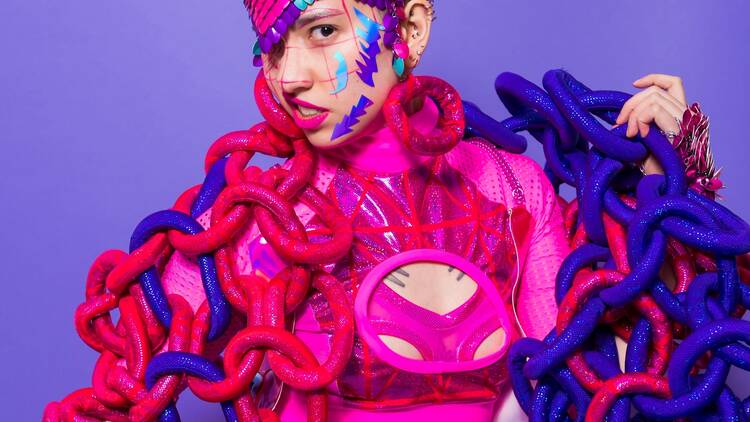 A model poses in an ostentatious pink and purple outfit on a purple background