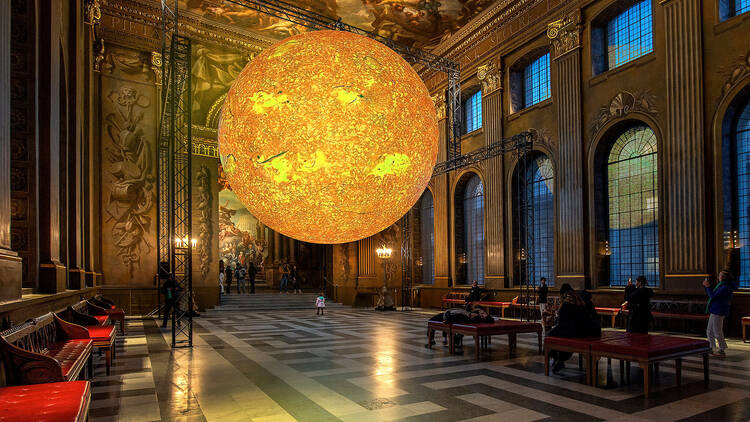 A large glowing yellow orb that looks like the sun is suspended in the centre of a grandly decorated Baroque hall with painted walls and ceilings,