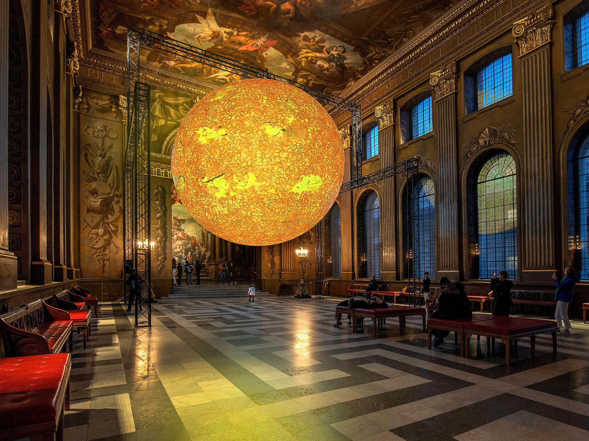 A large glowing yellow orb that looks like the sun is suspended in the centre of a grandly decorated Baroque hall with painted walls and ceilings,