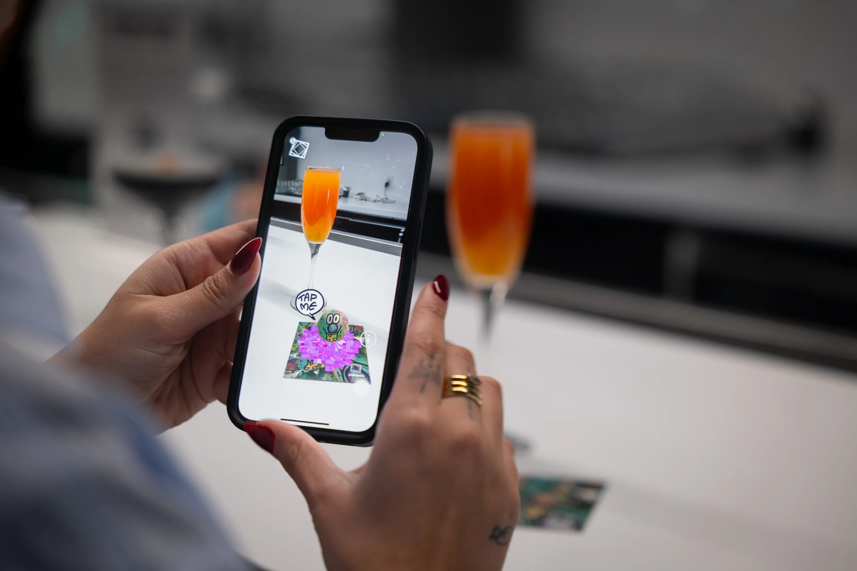 A person holds up a phone next to a cocktail, with a digital character popped up on the phone's screen.