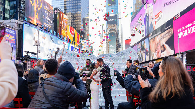 A couple gets married in Times Square, kissing as confetti falls around them.