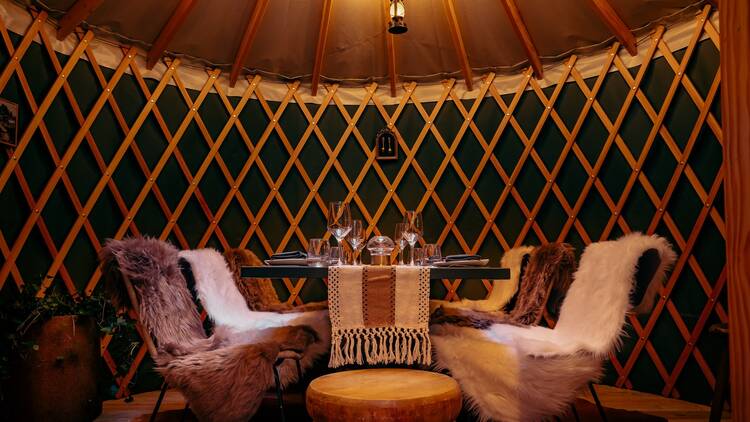 Four chairs covered in furs inside a cabin centered around a table