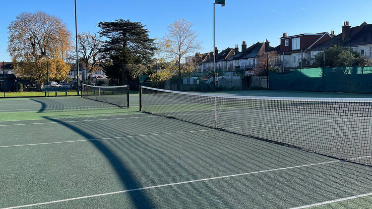 Two green tennis courts with long shadows of the nets cast across the courts. Houses overlooking the courts