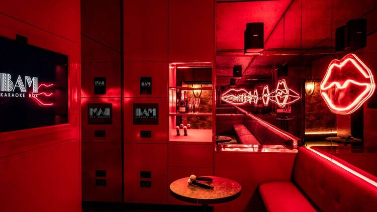 A karaoke booth with a TV screen, two microphones sat on a round table, and a banquette seat, bathed in red light with a red neon sign shaped like lips