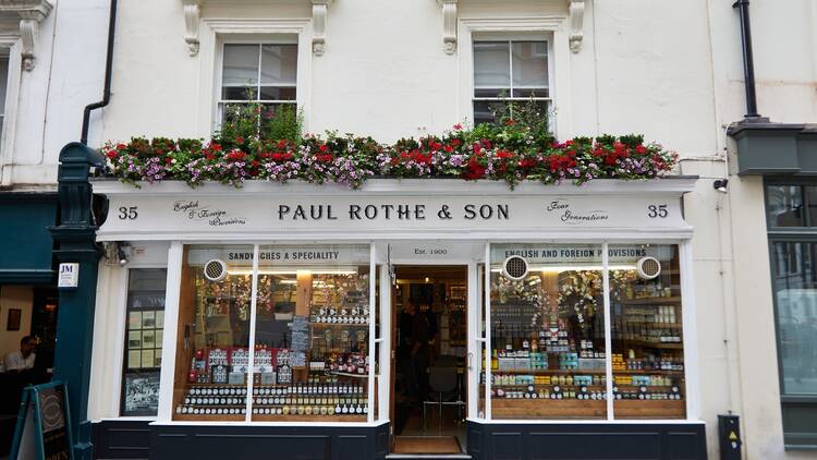 A shop exterior painted white, with 'Paul Rothe & Son' in black lettering outside. There are colourful flowers lining the guttering, and jars of condiments stacked high in the windows.