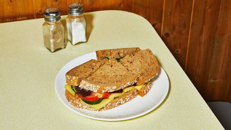 A well filled sandwich made with brown bread and cut into quarters sits on a white plate, on top of a yellow formica table. Salt and pepper shakers are to its left, and wood panelled walls in the background