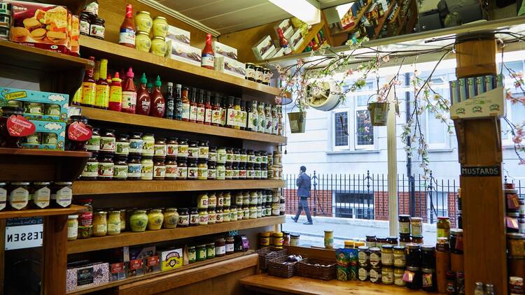 Rows of condiments including chilli sauces, jams and mustards sit on wooden shelves in front of a window looking out onto the street, illuminated by a flourescent strip light on the ceiling 