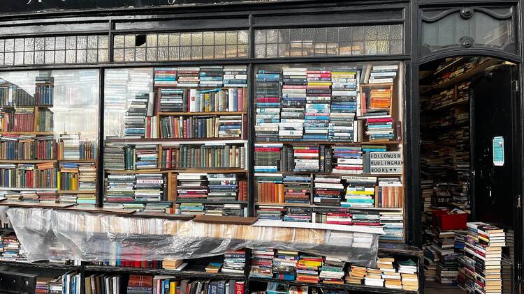 The exterior of Hurlingham Books on Fulham High Street, a black shopfront with three large windows piled high with disorderly stacks of books 