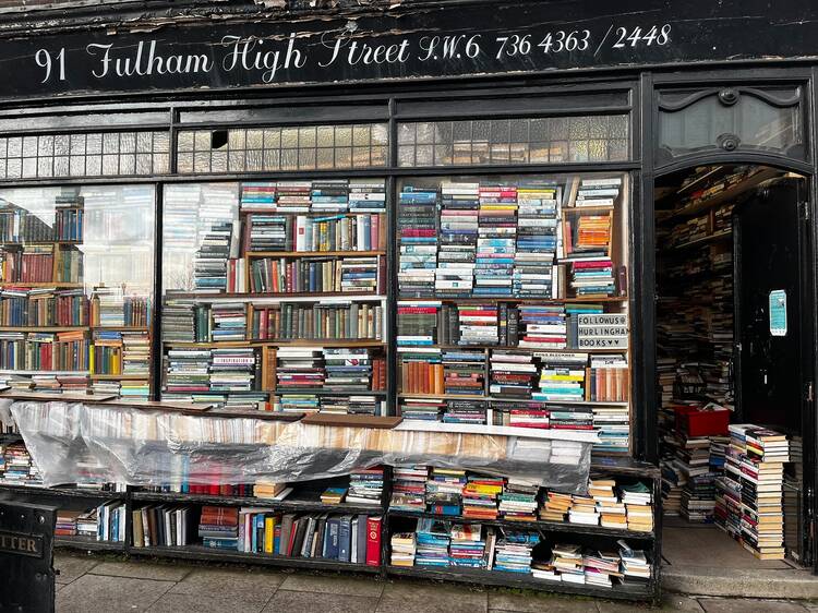 The exterior of Hurlingham Books on Fulham High Street, a black shopfront with three large windows piled high with disorderly stacks of books 