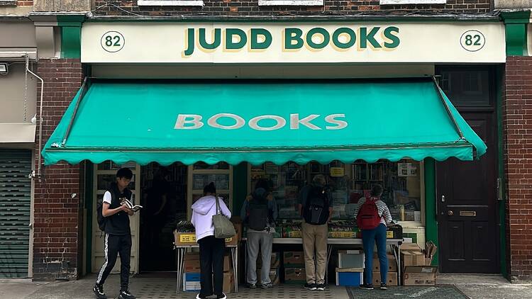 Exterior of Judd Books; the name of the shop appears in green capital letters on a cream background on the shopfront, with a mind green awning with the word 'books' in gold capitals obscuring sheltering shelves of books outside the front window.