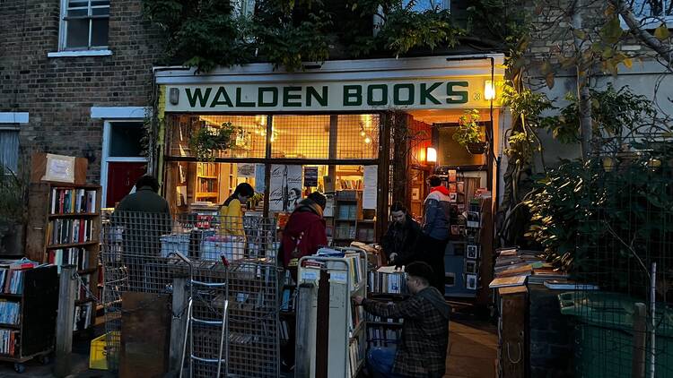 A small crowd of people browsing shelves of books on the pavement outside a small shopfront with Walden Books in green capital letters on the white shopfront. It is almost dark, and the shop is illuminated from inside by yellow light.