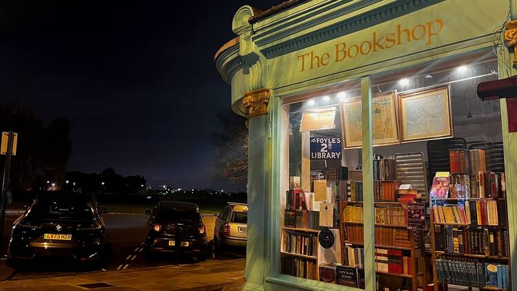 Exterior of the Bookshop on the Heath, which has a mint green exterior with gold serif font lettering saying 'The bookshop', viewed at night