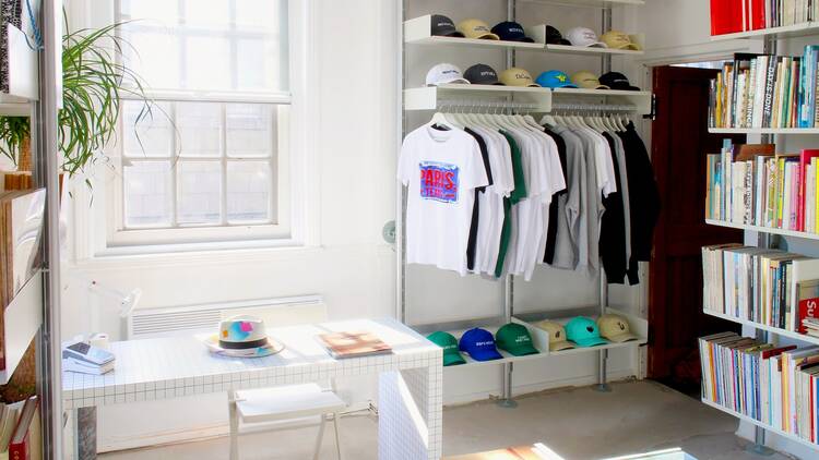 A brightly lit white-walled room in which several neatly arranged bookshelves stand, alongside shelves filled with colourful baseball caps, and a line of t-shirts hanging from a railing