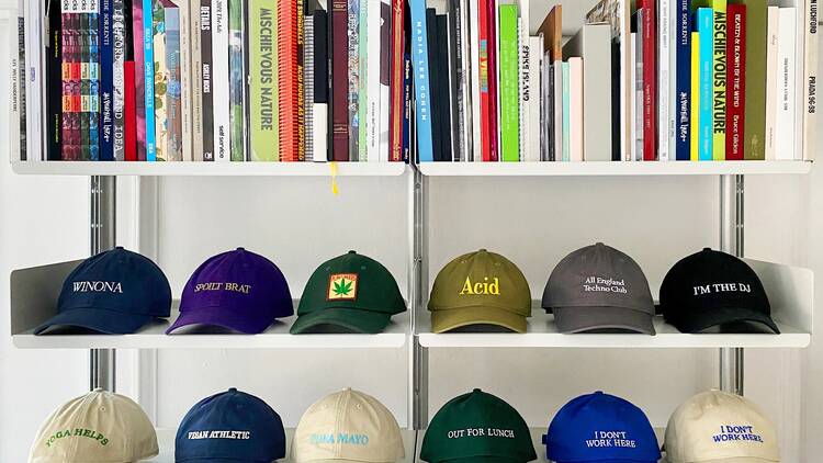 Twelve different coloured baseball caps with an array of slogans and designs sit on two shelves, with a shelf above these stacked with books