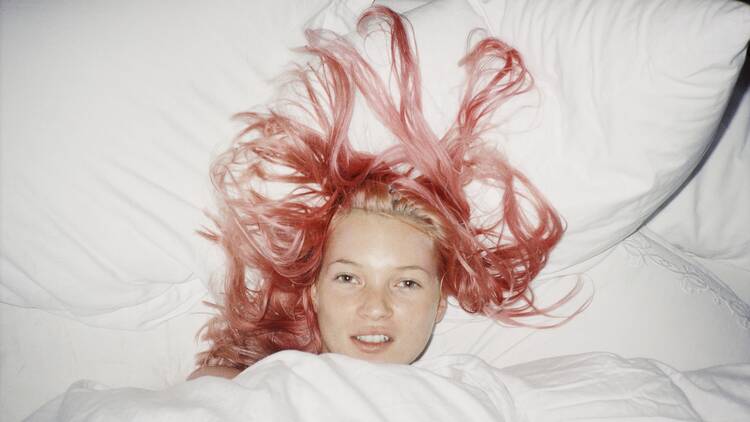 A pink-haired young Kate Moss tucked into a bed with white sheets, photographed by Juergen Teller, 1998