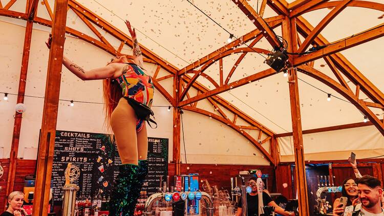 A drag queen stands on top of the bar in a tent at Between the Bridges