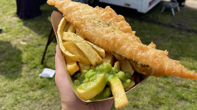 Hand holding a cardboard of battered fish, chips and mushy peas