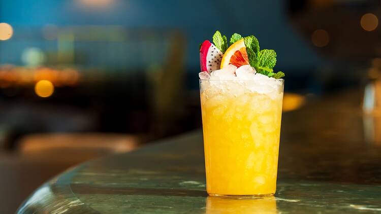 A glass containing crushed ice and bright orange drink, garnished with dragon fruit, blood orange and mint, sititng on a grene marble bar